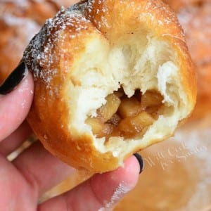 3 easy apple pie doughnuts on a decorative wooden plate with a white and red fall festive cloth in the background and a hand holding one doughnut that has a bite in it.