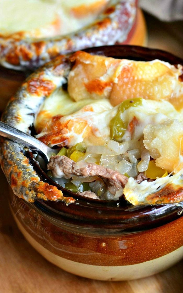 2 clay crocks filled with philly cheese steak soup on a wooden cutting board with a spoon resting inside of first crock as viewed close up