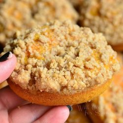 hand holding 1 pumpkin carrot cake streusel donut above a pile of stacked donuts on a wooden table