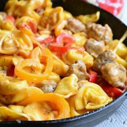 black skillet filled with sausage peppers and onion tortellini dish on a light blue cloth with a red and white cloth in the background as viewed close up