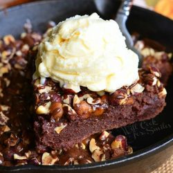 1 slice of brownie being held above black skillet filled with a small batch of pumpkin turtle brownie on a wooden table as viewed close up