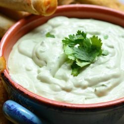 Avocado ranch dip in a blue and orange crock with taquitos leaning on crock