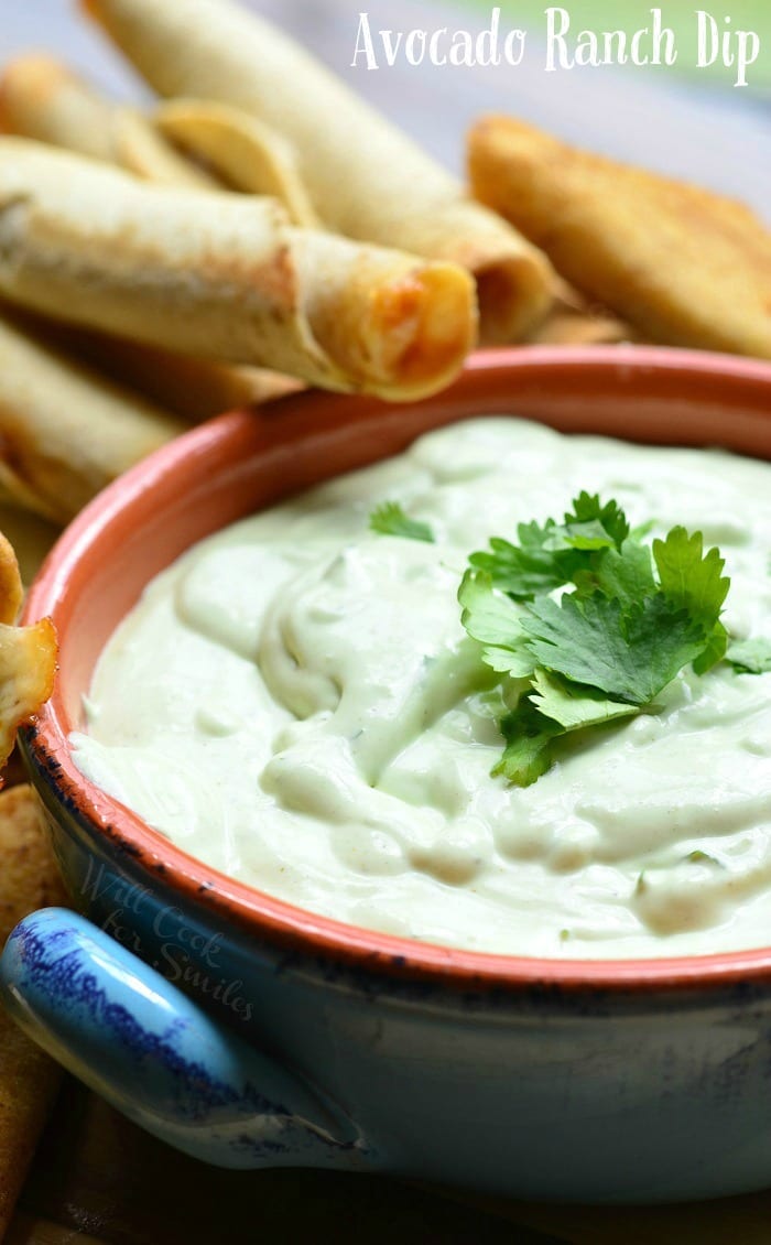 Avocado ranch dip in a blue and orange crock with taquitos leaning on crock