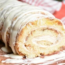 Close up view of cinnamon roll cake roll on a wooden board with icing drizzled across the top of the roll and a white and red cloth in the background