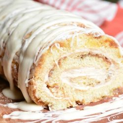 Close up view of cinnamon roll cake roll on a wooden board with icing drizzled across the top of the roll and a white and red cloth in the background