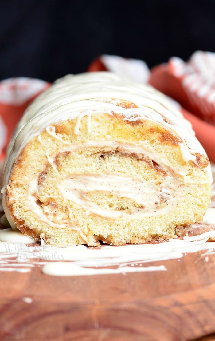 View from the front of cinnamon roll cake roll on a wooden board with icing drizzled across the top of the roll and a white and red cloth in the background