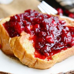 small round white plate with cranberry bliss french toast on a brown burlap placemat, a fork in the background to the right and several cranberries scattered around the plate