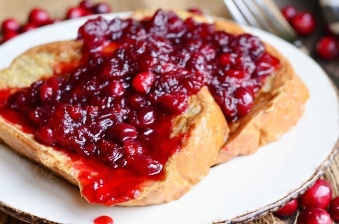A plate with French toast topped with a generous serving of red cranberry sauce. Some whole cranberries are scattered around the plate.