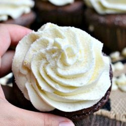 Double chocolate cupcakes with white chocolate cream cheese frosting on a white and black placemat with white chocolate chips scattered around the bottom of the cupcakes and a hand holding 1 cupcake in the foreground