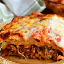 1 slice of Italian sausage lasagna on a light yellow colored plate on a wooden table with the rest of the lasagna in a clear baking dish in the background to the right