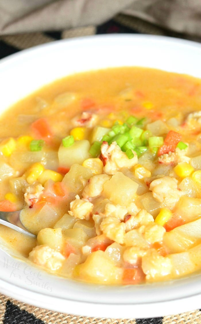 view from above of a white bowl filled with lobster potato and corn chowder as a spoon rests on the bowl and a brown cloth sits in the background