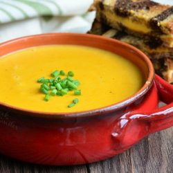 Red clay crock filled with pumpkin ale soup on a wooden table with a cut up grilled cheese in the background and a white and green cloth in the background to the left