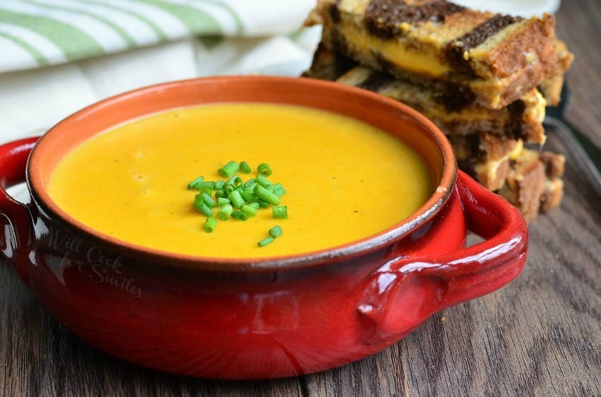 Pumpkin Ale Soup in a orange soup bowl with chives on top as a garnish and pumpernickel grilled cheese behind the bowl 