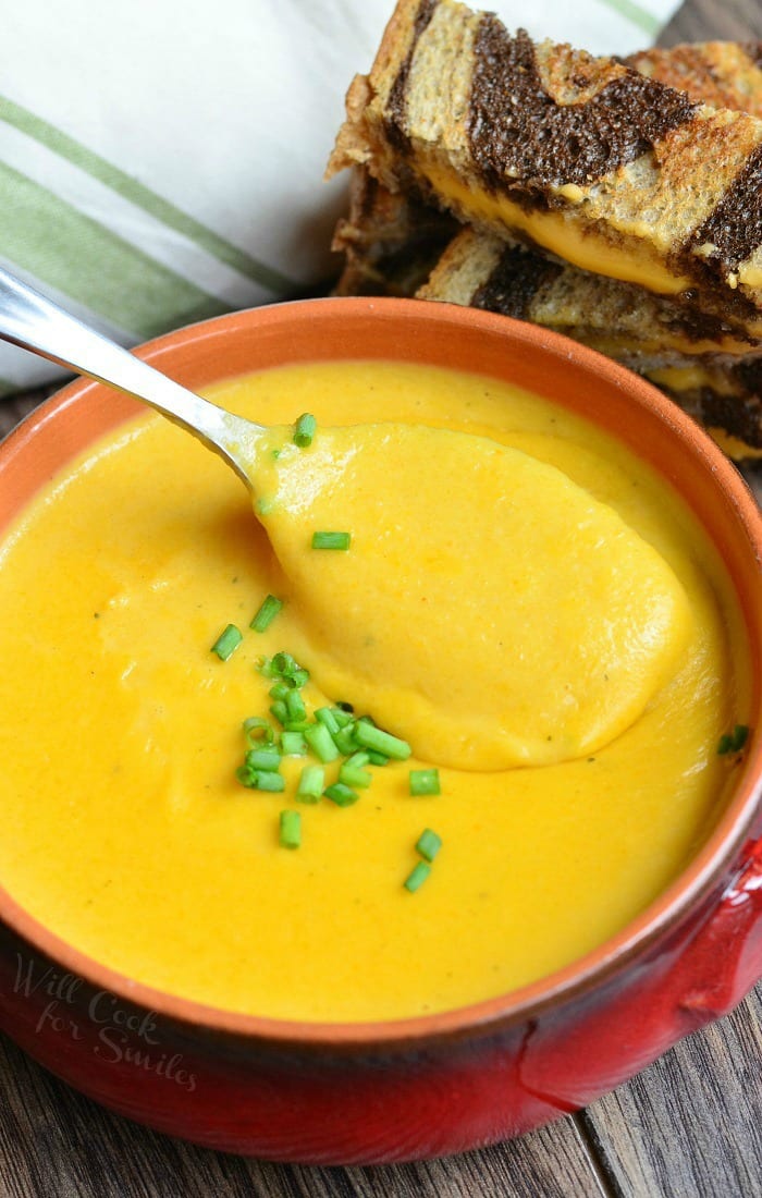 Pumpkin Ale Soup in a orange soup bowl with chives on top as a garnish and pumpernickel grilled cheese behind the bowl with a spoon