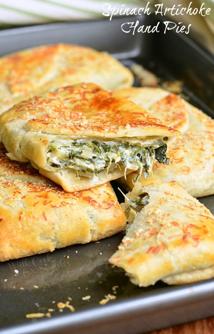Spinach Artichoke Hand Pies stacked up on a baking sheet with the top one cut in half 