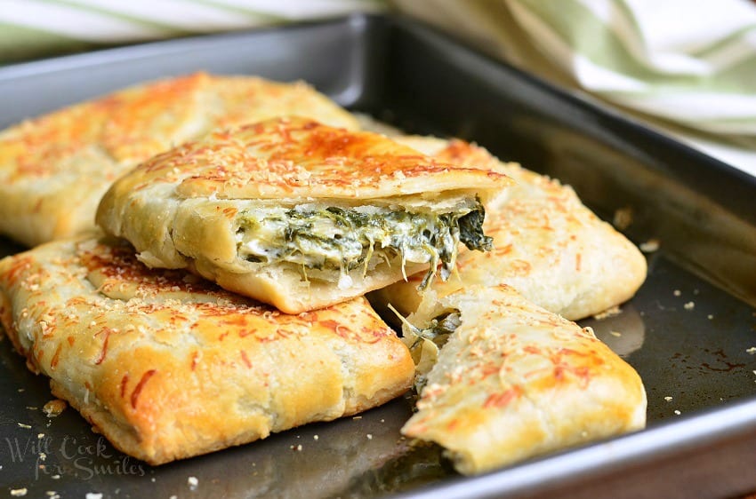 Spinach Artichoke Hand Pies stacked up on a baking sheet with the top one cut in half 