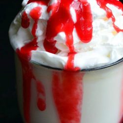 Clear glass coffee mug filled with white chocolate raspberry halloween hot chocolate on a black table with raspberry sauce splatter on table around base of glass viewed close up