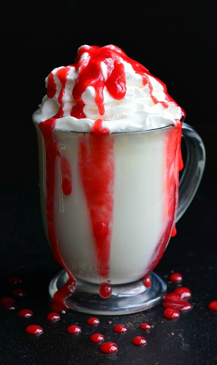 White Chocolate Raspberry Hot Chocolate with whip cream on top and red raspberry sauce in a clear glass mug with drops of raspberry sauce around the glass on the black table 