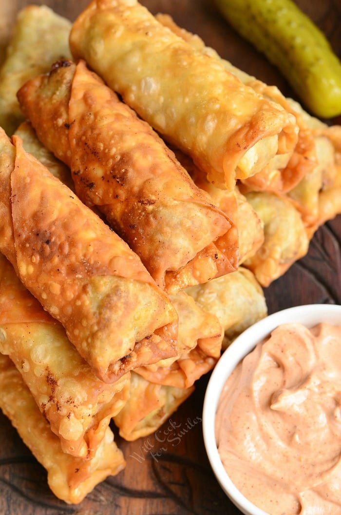 top view of cheeseburger Egg Rolls stacked on cutting board with bowl of sauce on the right side and a pickle at the top right corner 