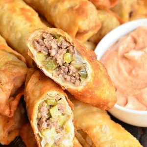 stacked cheeseburger eggrolls on a wooden tray with a small white bowl filled with dipping sauce and an eggroll cut in half and sitting on top of the stack of eggrolls
