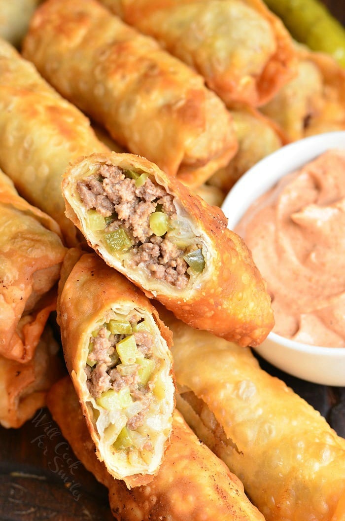 Cheeseburger Egg Rolls with one cut in half and the ground beef and pickles showing and a bowl of sauce to the right side 