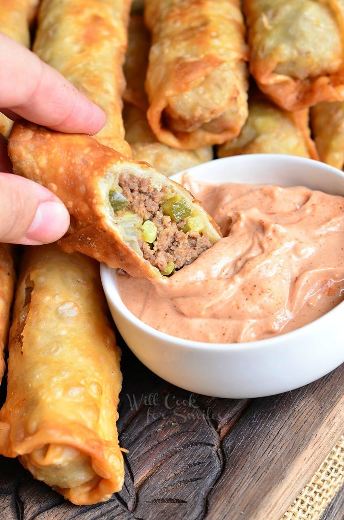 Cheeseburger Egg Rolls on a cutting board dipping on in sauce that is in white bowl 