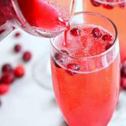 3 champagne glasses filled with cranberry pomegranate mimosas on a white table with cranberries scattered along the base of the glasses and more being poured into the glass in the foreground