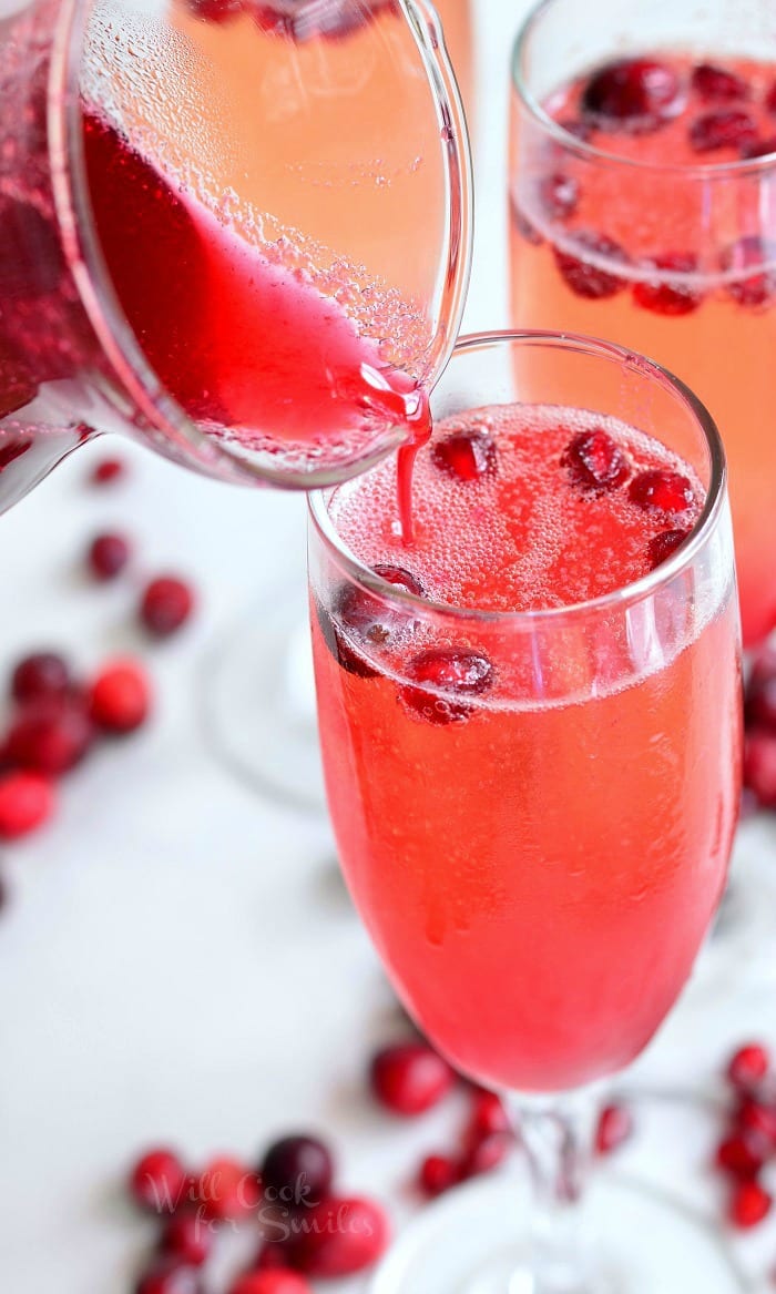 pouring Cranberry Pomegranate Champagne Cocktail in a champagne glass with Pomegranate and cranberries in the glass as garnish and around the bottom of the glass 
