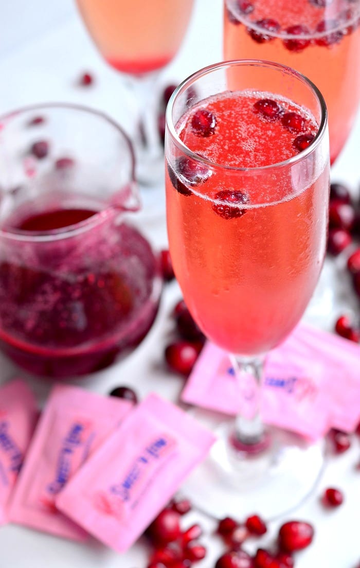 top view photo of Cranberry Pomegranate Champagne Cocktail in a champagne glass with Pomegranate and cranberries in the glass as garnish and around the bottom of the glass with packets of sweet and low 