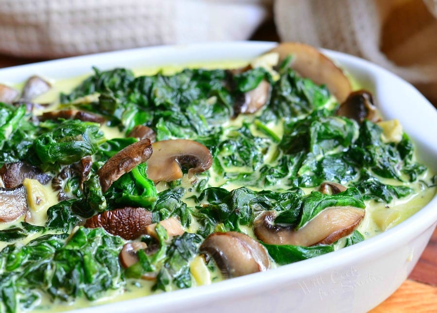 close up view of a white baking pan filled with creamed spinach and mushroom in white wine sauce on a wooden table with a white cloth behind