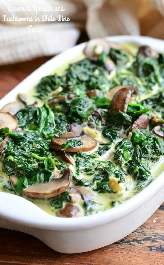 white baking pan filled with creamed spinach and mushroom in white wine sauce on a wooden table with a white cloth behind