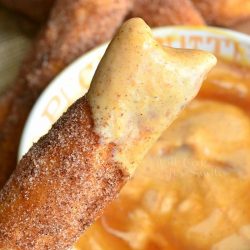 a tan bowl filled with pumpkin yogurt dip on wood cutting board with easy sugar doughnut sticks piled next to bowl and a hand holding 1 stick after being dipped in dip