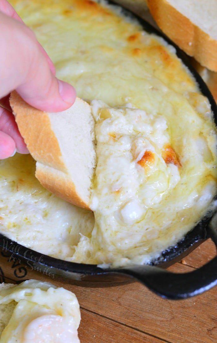 dipping a slice of french bread into Seafood & Cheese Dip that is a cast iron pan 