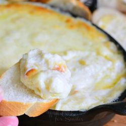 A hand hold a slice of bread with some dip over a black deep skillet filled with baked seafood cheese dip on a wooden table with sliced bread behind and to the right and a white cloth with black lettering in the background