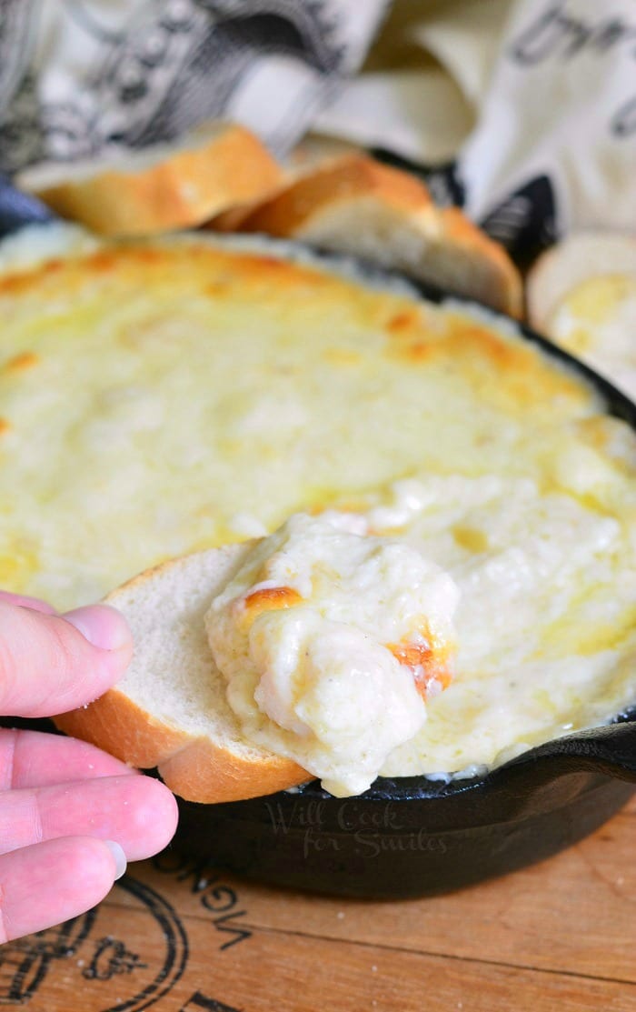 dipping a slice of french bread into Seafood & Cheese Dip that is a cast iron pan 