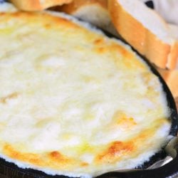 Black deep skillet filled with baked seafood cheese dip on a wooden table with sliced bread behind and to the right and a white cloth with black lettering in the background