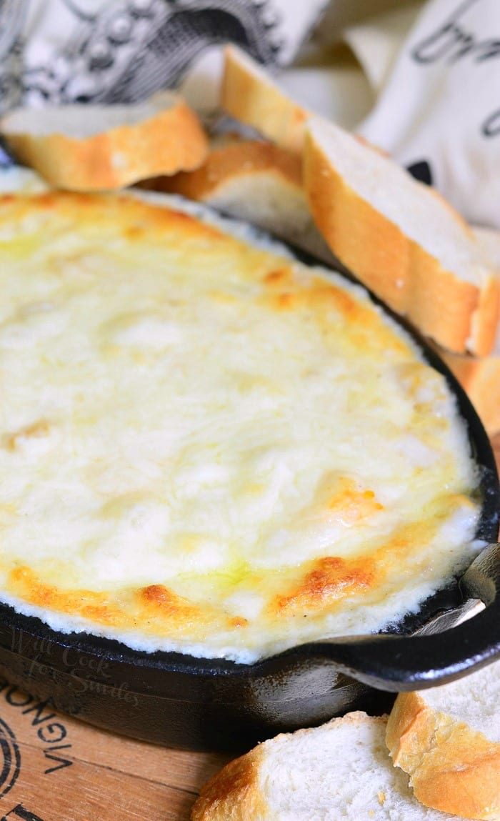 Black deep skillet filled with baked seafood cheese dip on a wooden table with sliced bread behind and to the right and a white cloth with black lettering in the background