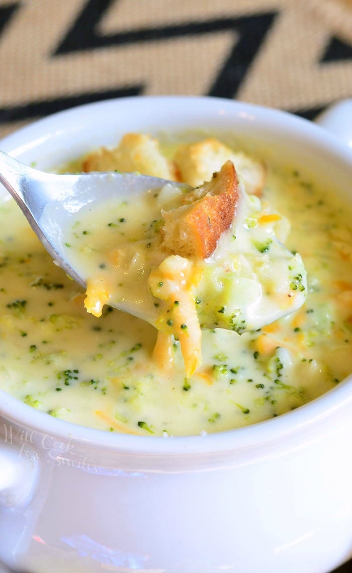 White crock filled with asiago cheese soup on a yellow plate with cut bread croutons scattered around the bowl placed on the plate with a spoon pulling out one portion of soup