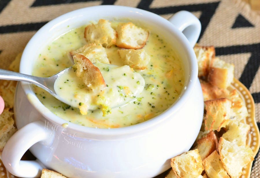 Asiago Broccoli Cheese Soup in a white soup bowl with cut up pieces of crusty bread on top and around the bottom of the bowl