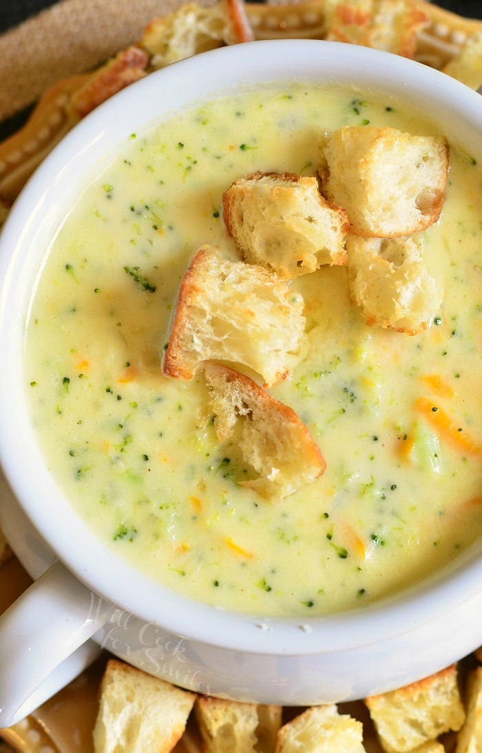 Top view of Asiago Broccoli Cheese Soup in a white soup bowl with cut up pieces of crusty bread on top and around the bottom of the bowl 