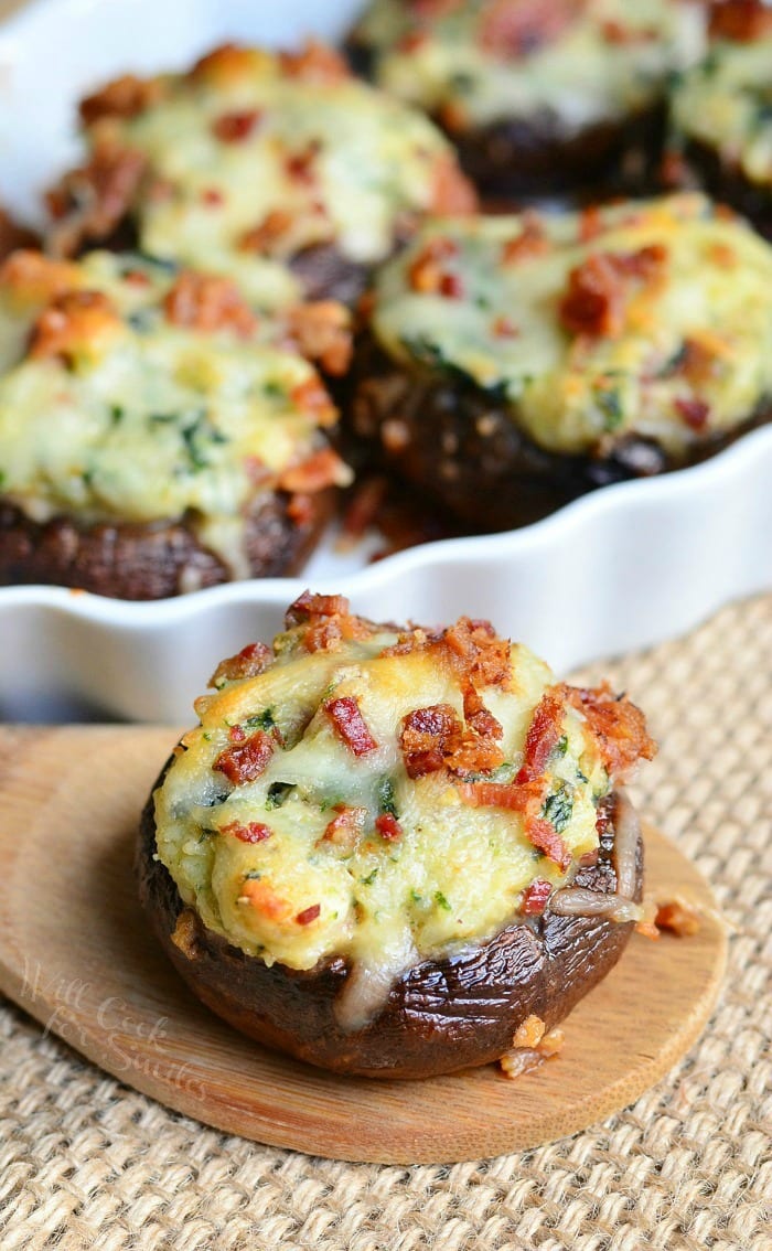 Decorative oval platter filled with bacon spinach and four cheese stuffed mushrooms in the background with 1 stuffed mushroom on a wooden spoon in the foreground.