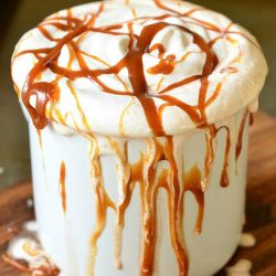 Metal dessert mug filled with caramel marshmallow hot chocolate on a wooden table covered in caramel drizzle