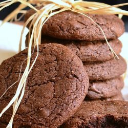 2 chocolate chocolate chip gingersnap cookies in front of additional cookies stacked behind on a white cloth while all sit on a wooden table