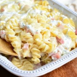Close up view of White baking dish filled with creamy chicken cordon bleu pasta casserole on a wooden table with a wooden serving spoon pulling out one helping of the dish