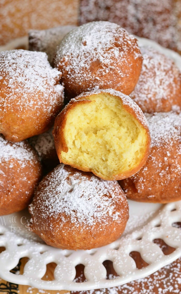 Easy Ricotta Doughnuts piled on a decorative round white plate with powdered sugar topping the doughnuts with one doughnut bit into