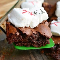 A small green spatula holding 1 gooey peppermint marshmellow brownie above a wooden table as a glass baking dish and additional brownies rest in the background