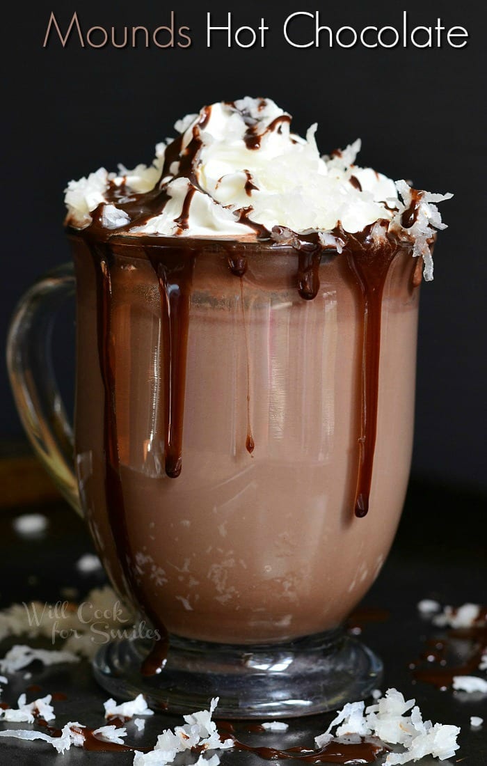 Mounds Hot Chocolate in a clear glass mug with whip cream and coconut on top and chocolate sauce on top and spilling over the cup 