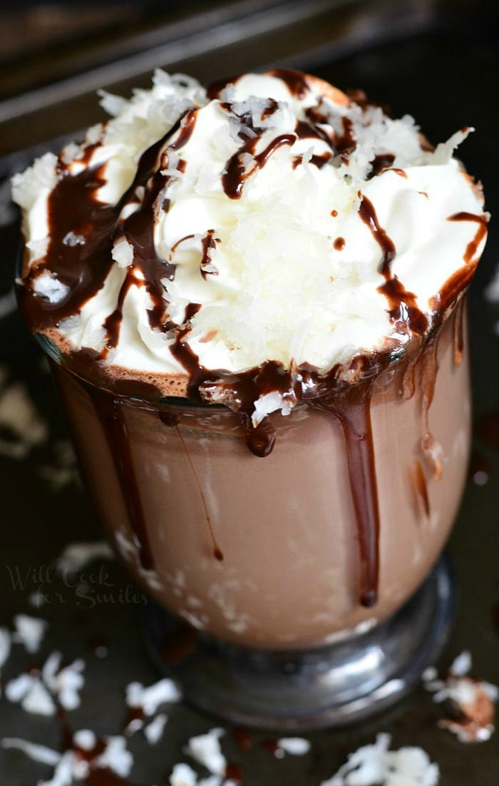 Hot Chocolate in a clear glass with whipped cream, chocolate sauce, and shredded coconut on top  