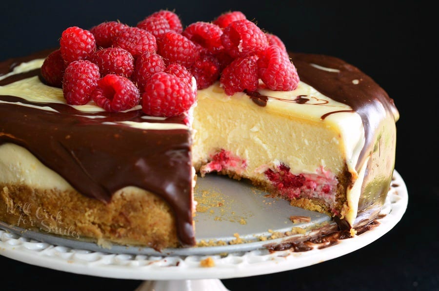 Double Chocolate Ganache and Raspberry Cheesecake with raspberries on top and a piece missing on a cake stand 