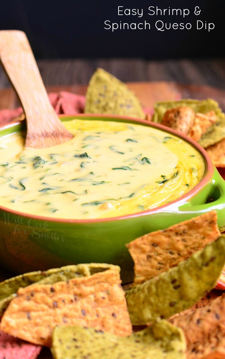 Shrimp and Spinach Queso Dip in a green bowl with a wooden spoon in it and tortilla chips around it 